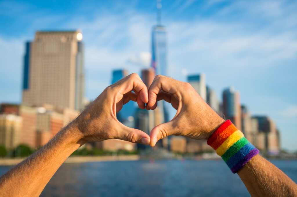 Hands in heart formation with rainbow armband and NYC skyline in the background (Photo Credit: shutterstock)